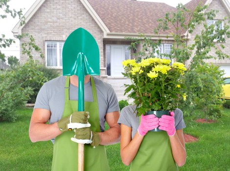 In a suburban garden setting, two individuals are standing on a well-maintained lawn in front of a house with a light brick facade, white-framed windows, and a sloped tiled roof, surrounded by green bushes and small trees. One person, dressed in gardening gloves, an apron, and holding a green trowel, is wearing a bright green gardening helmet for safety. The other, wearing pink gloves, holds a large pot filled with yellow flowering plants, possibly chrysanthemums or similar, obscuring their face. The garden features a lush, evenly mowed grass area, with a paved pathway partially visible, and a flower bed along the houseâ€™s exterior. The weather appears bright and clear, with natural daylight illuminating the scene, emphasizing the vibrant colors of the plants and safety gear. This image reflects professional gardening and outdoor maintenance activities typical for a residential garden in Uxbridge, supporting services offered by Gardening Services Uxbridge.