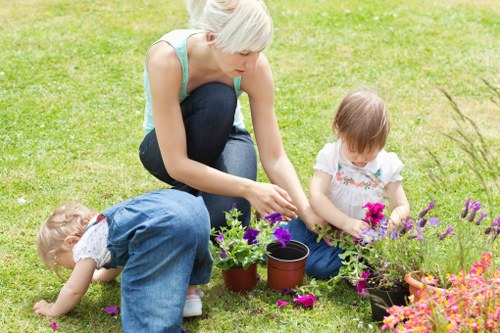 A woman with short blonde hair, wearing a light blue sleeveless top and dark jeans, is kneeling on a lush green lawn, engaging in gardening activity with two young children. She is tending to potted plants, one of which contains purple and green foliage, while the children, a girl in a white dress with floral patterns and a boy in a light-colored shirt and blue overalls, are actively involved in planting or watering. The garden features a well-maintained grassy area, flower beds with an assortment of colorful blooms, and a backdrop of trees and shrubs. Sunlight illuminates the scene, indicating a bright day with clear weather. This outdoor space, typical of residential gardens in Uxbridge, demonstrates a neat layout with defined planting areas and tidy grass, suitable for family gardening activities. The scene underscores professional gardening support to maintain such vibrant and inviting outdoor spaces in the local area, highlighting the importance of regular lawn care, flower bed maintenance, and general garden upkeep offered by Gardening Services Uxbridge.
