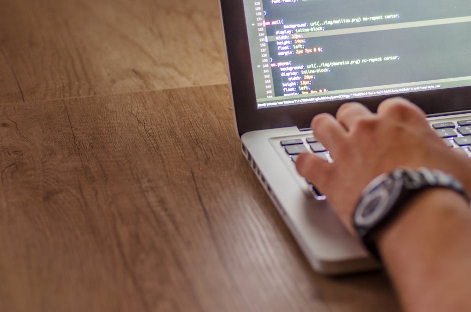 A person's hand resting on a silver laptop keyboard with visible lines of code on the screen, placed on a wooden surface with a natural grain texture. The individual is wearing a patterned wristwatch 