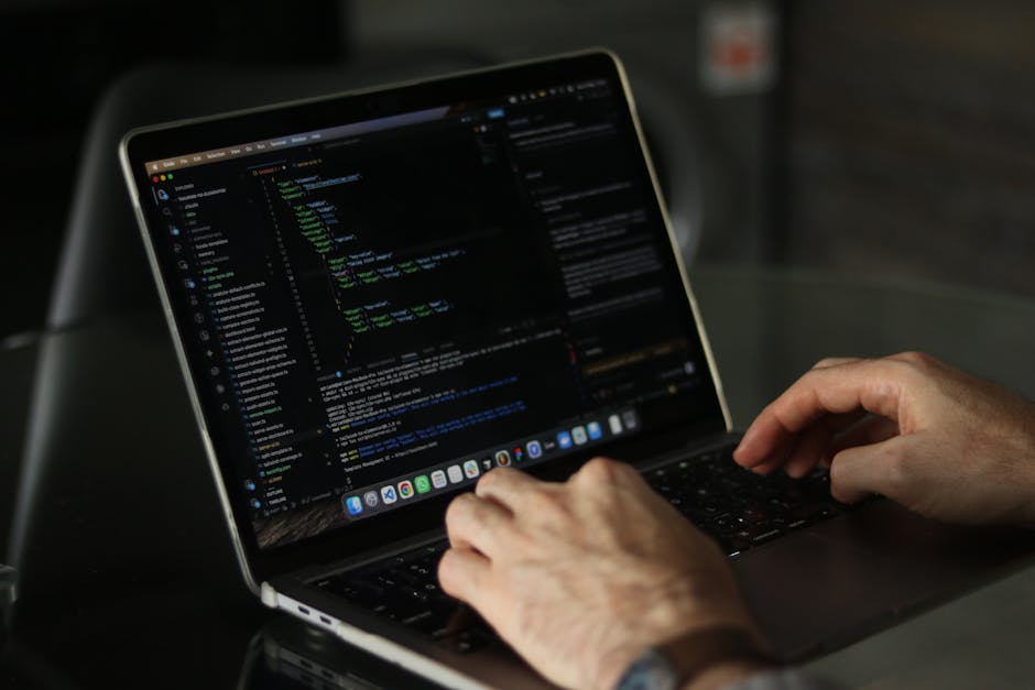 A person's hand resting on a silver laptop keyboard with visible lines of code on the screen, placed on a wooden surface with a natural grain texture. The individual is wearing a patterned wristwatch 