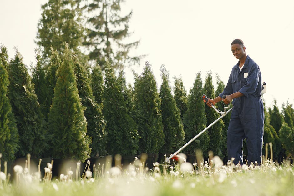 A person dressed in protective outdoor work clothing, including boots and orange high-visibility trousers, using a grass trimmer or line trimmer to cut and clear overgrown grass and weeds in a garden